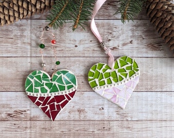 May include: Two heart-shaped mosaic ornaments with green and red, and green and pink designs, hang from a pink ribbon and a wire with beads. A pine branch and pine cone are in the background, set against a wood-grain surface.