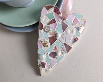 May include: A mosaic heart ornament with a pink ribbon for hanging. The heart is made of various colored tiles, including pink, white, and brown, with floral and button accents. A stack of blue-green teacups and saucers are in the background.