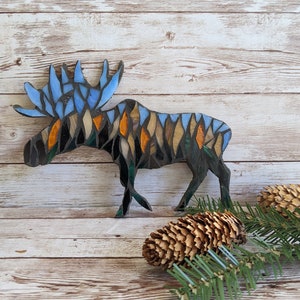 May include: A mosaic moose sculpture with blue, orange, and brown accents. The moose has large antlers and is set against a wooden background. Two pine cones and a sprig of evergreen are in the foreground.