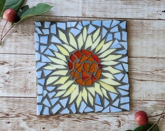 May include: Square mosaic art piece featuring a sunflower design. The center of the flower is made of orange and red glass pieces, surrounded by yellow petals. The background is light blue, with a dark gray border. The artwork is on a wooden surface.