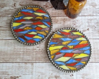 May include: Two circular coasters featuring a mosaic pattern of multicoloured glass shards in red, yellow, blue, and purple, set in grey grout with a beaded metal border. Behind them sit an amber glass bottle and a dark red glass goblet.