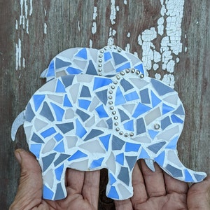 K&ouml;nnte beinhalten: Zwei Mosaik-Elefantenfiguren. Die Elefanten bestehen aus blauen, grauen und wei&szlig;en Glasst&uuml;cken. Die Elefanten haben silberfarbene Ohren und R&uuml;ssel. Die Elefanten haben eine Reihe silberfarbener Perlen auf dem R&uuml;cken.
