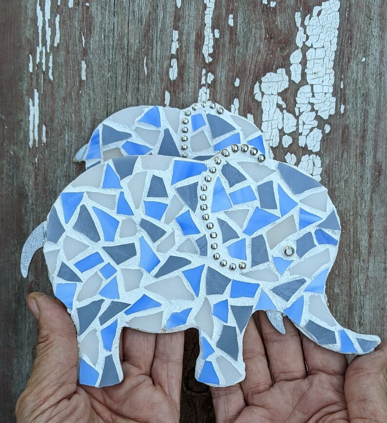K&ouml;nnte beinhalten: Zwei Mosaik-Elefantenfiguren. Die Elefanten bestehen aus blauen, grauen und wei&szlig;en Glasst&uuml;cken. Die Elefanten haben silberfarbene Ohren und R&uuml;ssel. Die Elefanten haben eine Reihe silberfarbener Perlen auf dem R&uuml;cken.