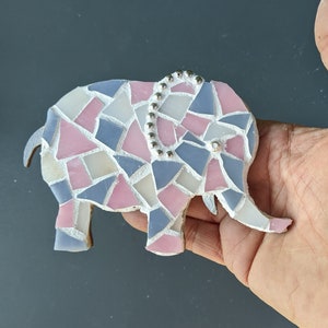 K&ouml;nnte beinhalten: Eine Mosaik-Elefantenfigur aus pastellrosa, blauen und wei&szlig;en Glasst&uuml;cken. Der Elefant hat eine dekorative Reihe silberner Perlen auf dem R&uuml;cken und eine einzelne Perle als Auge. Die Figur wird in einer Hand gehalten.