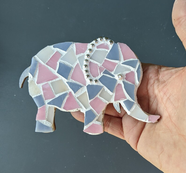 K&ouml;nnte beinhalten: Eine Mosaik-Elefantenfigur aus pastellrosa, blauen und wei&szlig;en Glasst&uuml;cken. Der Elefant hat eine dekorative Reihe silberner Perlen auf dem R&uuml;cken und eine einzelne Perle als Auge. Die Figur wird in einer Hand gehalten.