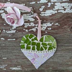 May include: A heart-shaped mosaic ornament with green and pink glass pieces. The ornament is adorned with silver-colored studs and a pink ribbon for hanging. A ceramic rose is next to the ornament.