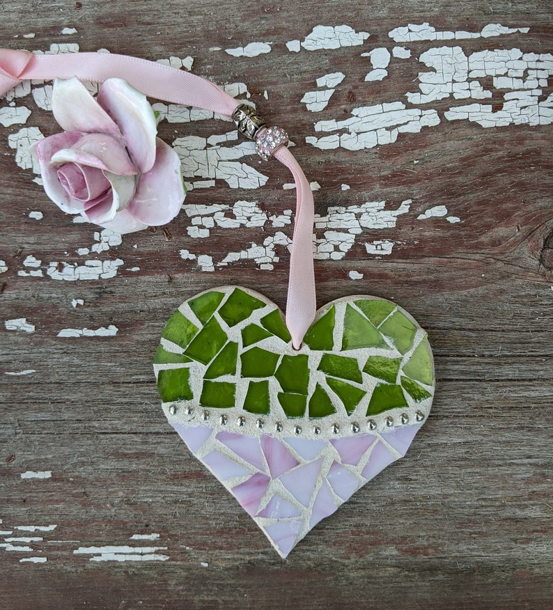 May include: A heart-shaped mosaic ornament with green and pink glass pieces. The ornament is adorned with silver-colored studs and a pink ribbon for hanging. A ceramic rose is next to the ornament.