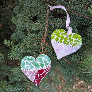 May include: Two heart-shaped Christmas ornaments with mosaic designs hang from a pine tree branch. One heart features green, pink, and red tiles, while the other has green and pink tiles. Both have a ribbon for hanging.