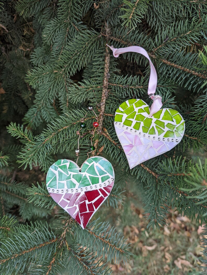 May include: Two heart-shaped Christmas ornaments with mosaic designs hang from a pine tree branch. One heart features green, pink, and red tiles, while the other has green and pink tiles. Both have a ribbon for hanging.