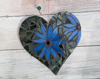 May include: A heart-shaped mosaic wall hanging with blue flower designs and green and blue glass pieces. The mosaic is suspended by copper wire and adorned with green and blue beads. The background is a light wood surface.
