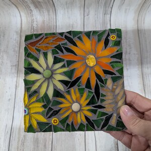 May include: A square mosaic art piece featuring yellow and orange flowers with green leaves and a dark gray background. The mosaic is made of small, colorful glass pieces. The piece is held by a hand.