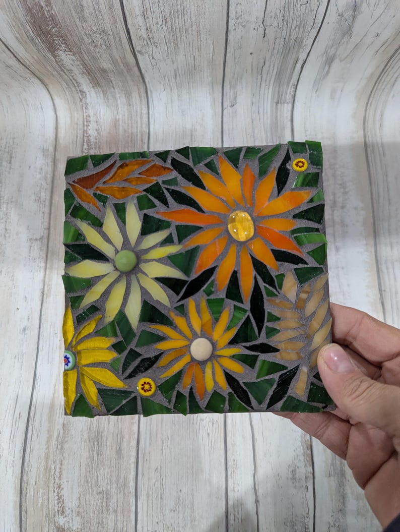 May include: A square mosaic art piece featuring yellow and orange flowers with green leaves and a dark gray background. The mosaic is made of small, colorful glass pieces. The piece is held by a hand.