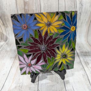 May include: A square mosaic art piece featuring colorful flowers. The flowers are in shades of blue, yellow, pink, and burgundy, with green leaves. The mosaic is set on a gray background and displayed on a black stand.