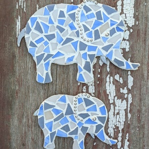 K&ouml;nnte beinhalten: Zwei Mosaik-Elefanten-Wandkunstwerke. Die Elefanten bestehen aus blauen, grauen und wei&szlig;en Glasst&uuml;cken. Der gr&ouml;&szlig;ere Elefant hat eine Perlenkette und der kleinere auch. Der Hintergrund ist eine verwitterte, rotbraune Holzoberfl&auml;che.
