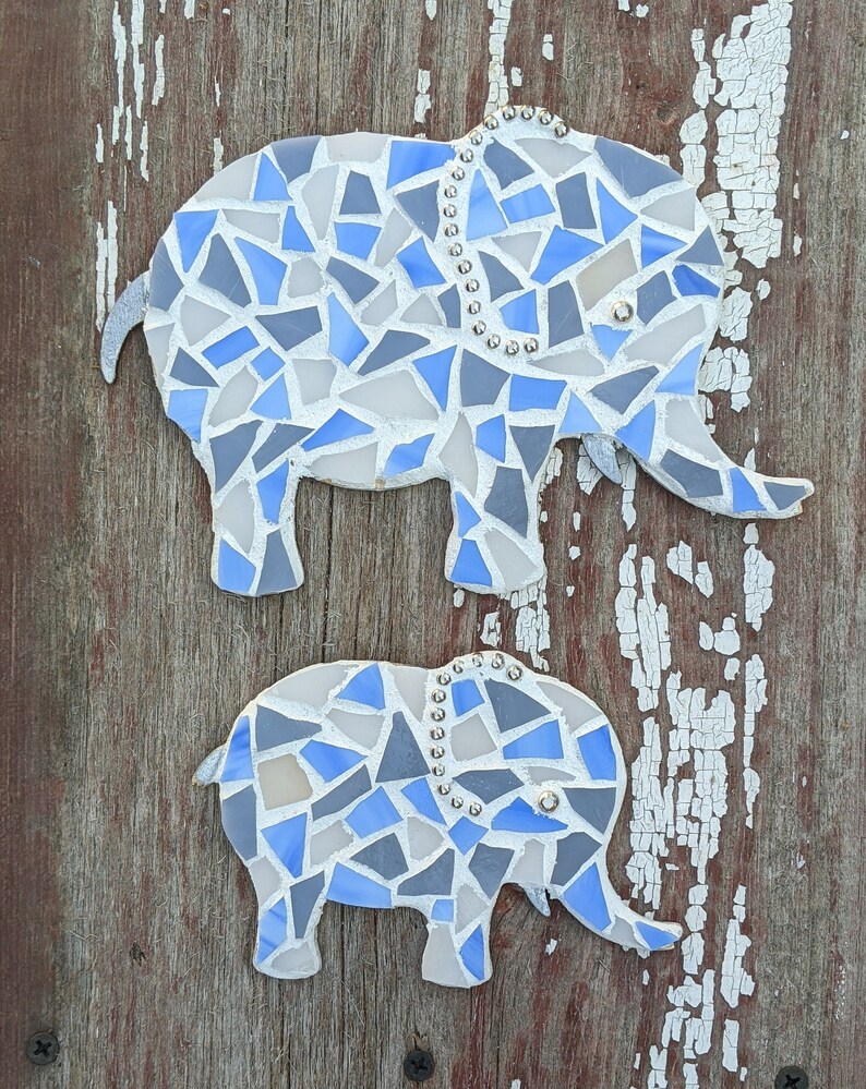 K&ouml;nnte beinhalten: Zwei Mosaik-Elefanten-Wandkunstwerke. Die Elefanten bestehen aus blauen, grauen und wei&szlig;en Glasst&uuml;cken. Der gr&ouml;&szlig;ere Elefant hat eine Perlenkette und der kleinere auch. Der Hintergrund ist eine verwitterte, rotbraune Holzoberfl&auml;che.