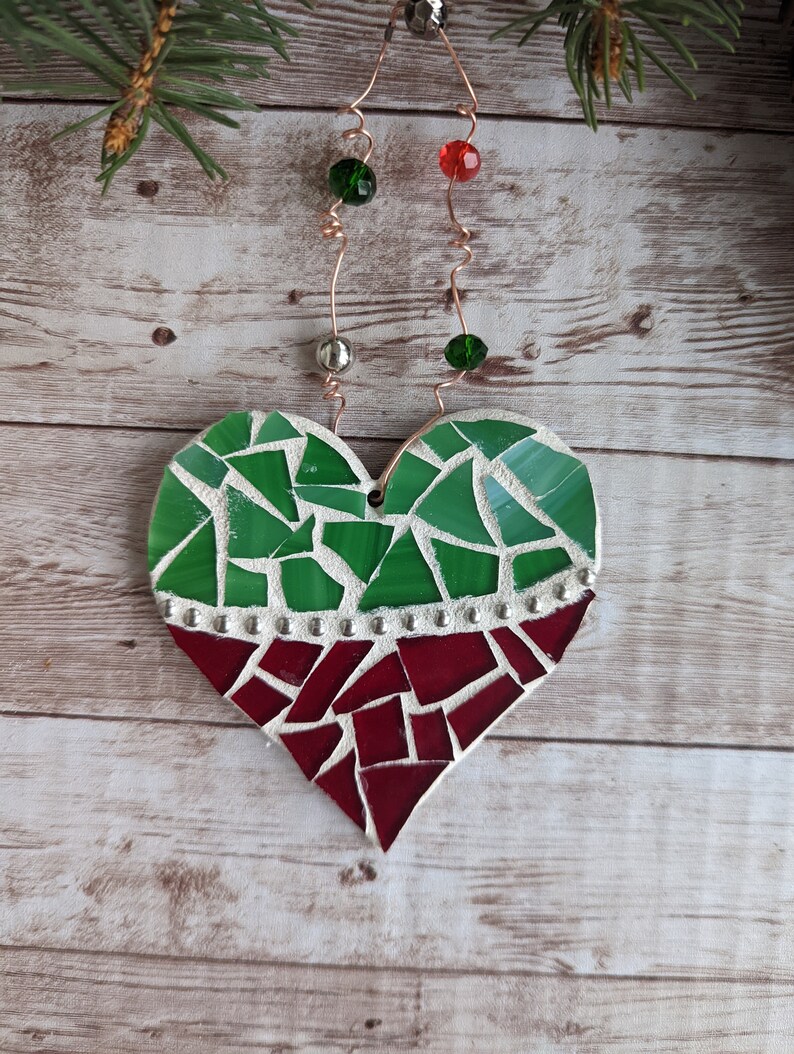 May include: A mosaic heart ornament with green and red glass pieces. The top half is green, the bottom red, separated by a row of silver studs. It hangs from copper wire with green and red beads, and silver beads.