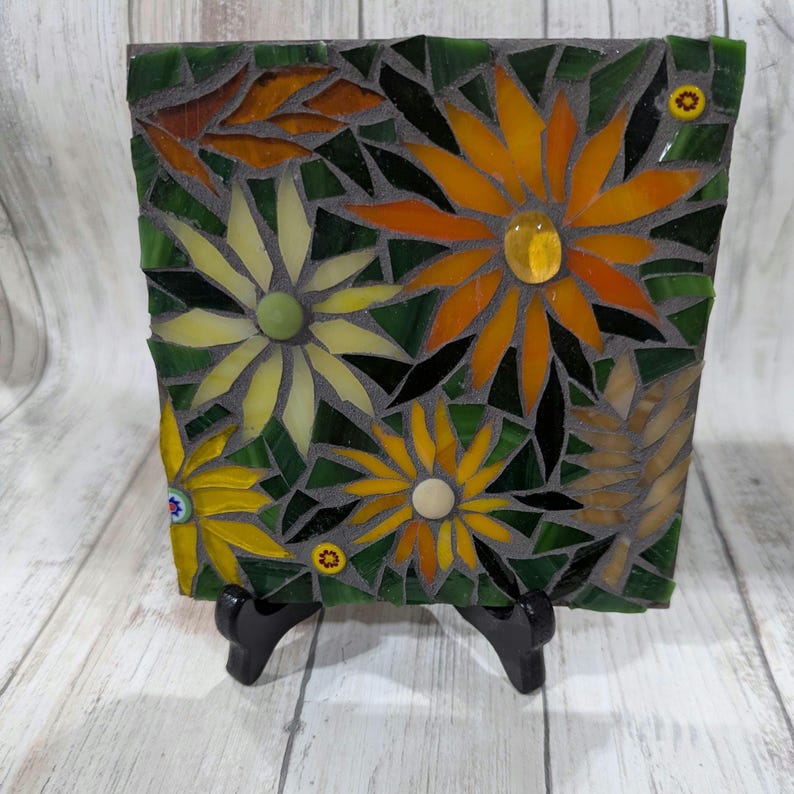 May include: A square mosaic art piece featuring a floral design. The mosaic is composed of various colored glass pieces, including yellow, orange, green, and brown. The flowers are set against a green background, with a dark gray grout. The piece is displayed on a black easel.