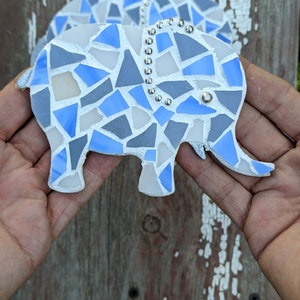 K&ouml;nnte beinhalten: Zwei Elefanten-Ornamente aus Mosaik. Jeder Elefant besteht aus blauen, grauen und wei&szlig;en Glasst&uuml;cken. Silberfarbene Nieten verzieren die R&uuml;cken der Elefanten. Die Ornamente werden vor einem verwitterten Holzgrund gehalten.