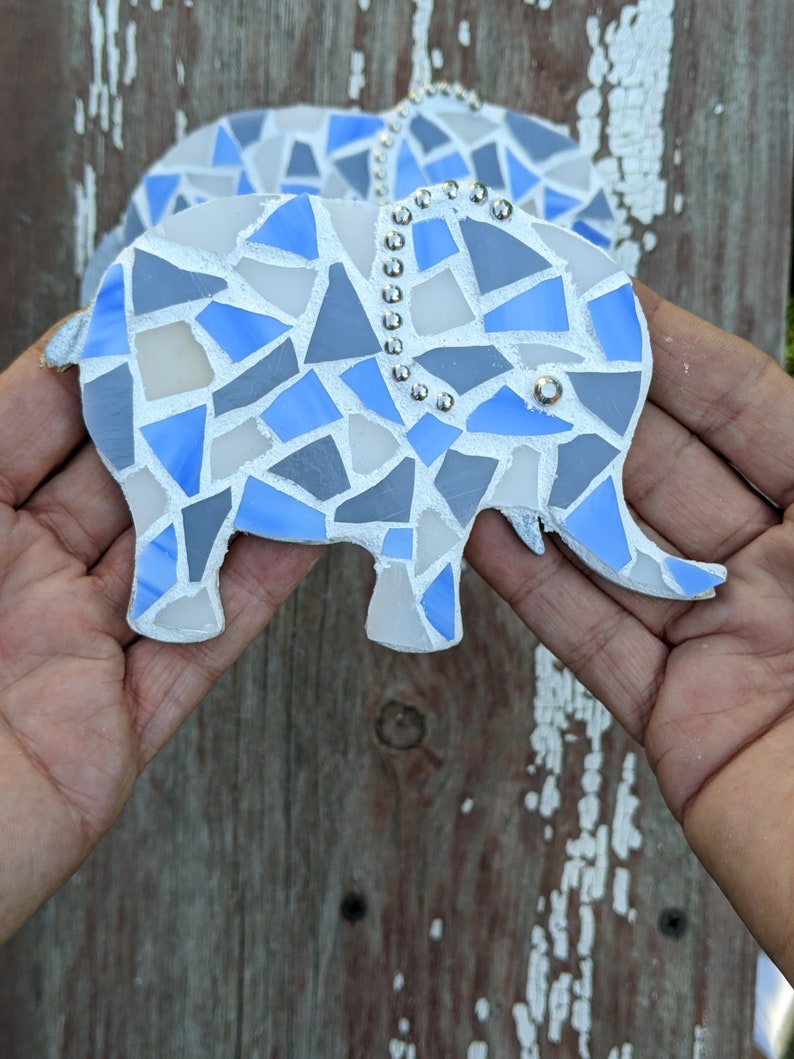 K&ouml;nnte beinhalten: Zwei Elefanten-Ornamente aus Mosaik. Jeder Elefant besteht aus blauen, grauen und wei&szlig;en Glasst&uuml;cken. Silberfarbene Nieten verzieren die R&uuml;cken der Elefanten. Die Ornamente werden vor einem verwitterten Holzgrund gehalten.