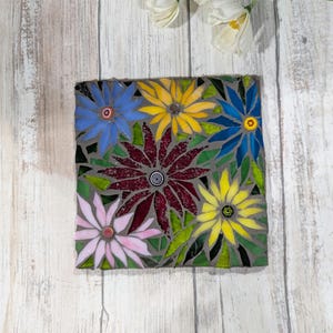 May include: A square mosaic featuring colorful flowers. The flowers are in shades of blue, yellow, pink, and burgundy, with green leaves. The mosaic is set against a gray background.