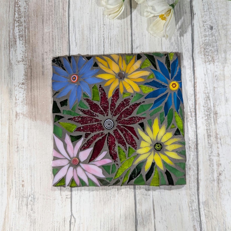 May include: A square mosaic featuring colorful flowers. The flowers are in shades of blue, yellow, pink, and burgundy, with green leaves. The mosaic is set against a gray background.