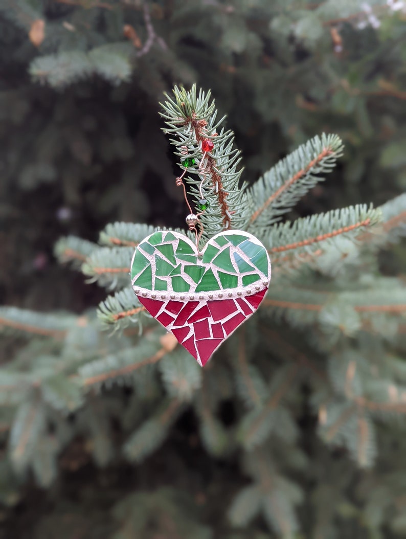 May include: A mosaic heart ornament with green and red glass pieces, hanging from a pine branch. The heart is divided into two sections, with a silver border. The ornament is decorated with small red and green beads.