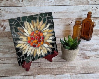 May include: A mosaic tile art piece featuring a sunflower design. The mosaic has a dark green background with a sunflower in shades of yellow, orange, and red. The artwork is displayed on a red easel, with brown glass bottles and a small potted succulent nearby.