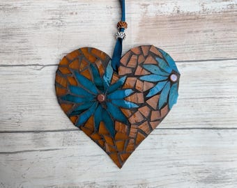 May include: A mosaic heart-shaped ornament with a teal ribbon for hanging. The heart is made of brown and teal glass pieces, with teal flower designs. A decorative bead accents the ribbon.