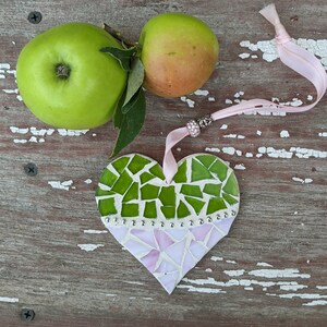 May include: A handmade mosaic heart ornament with green and pink glass pieces, accented with silver beads and a pink ribbon. Two green apples with a stem and leaves are also in the image, set against a rustic wooden background.