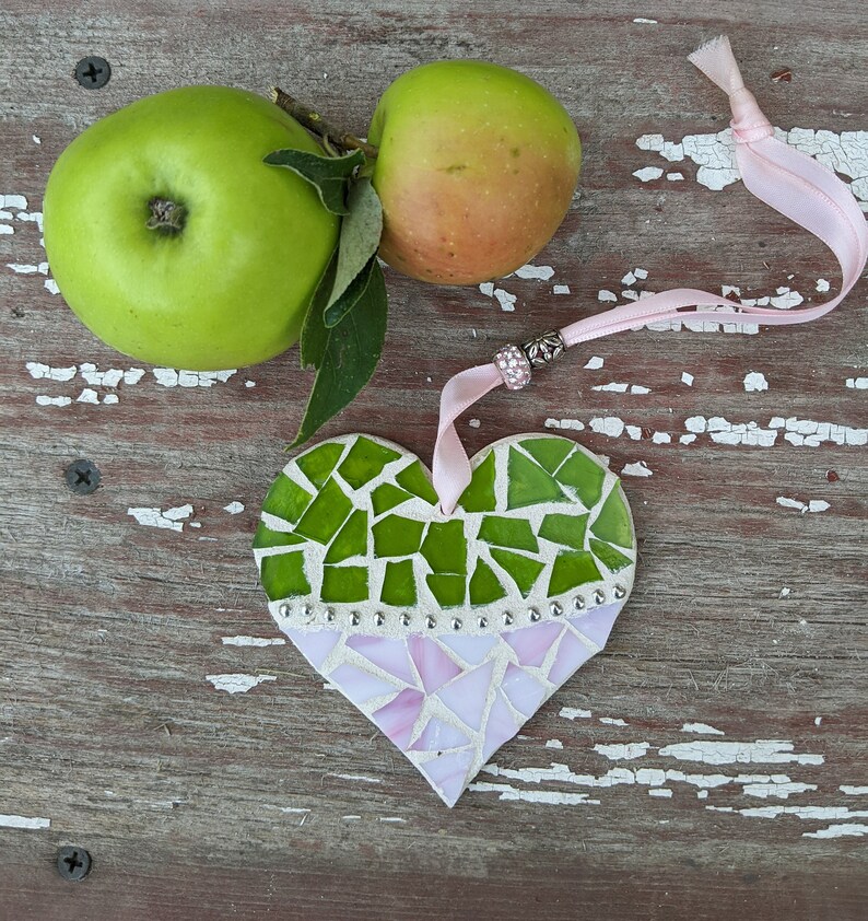 May include: A handmade mosaic heart ornament with green and pink glass pieces, accented with silver beads and a pink ribbon. Two green apples with a stem and leaves are also in the image, set against a rustic wooden background.
