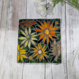 May include: Square mosaic tile featuring a floral design. The mosaic depicts yellow and orange flowers with green leaves and a gray background. The flowers have a detailed, textured appearance, and the tile is set against a light wood surface.