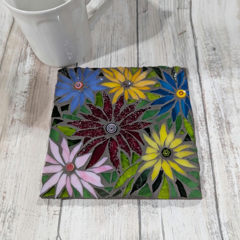 May include: Square mosaic coaster featuring a floral design. The coaster has a gray background with colorful flowers in shades of blue, yellow, pink, and burgundy. Green leaves are interspersed among the flowers. A white mug is in the background.