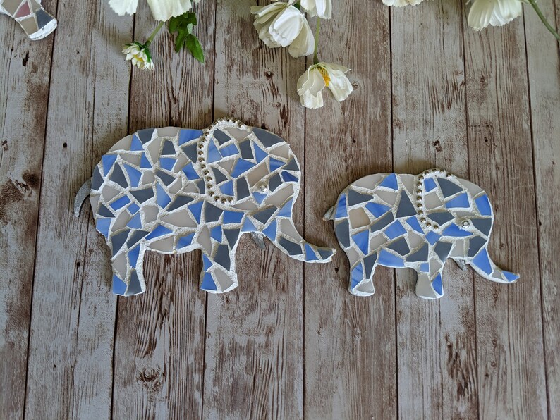 K&ouml;nnte beinhalten: Zwei Mosaik-Elefantenfiguren in Blau-, Grau- und Wei&szlig;t&ouml;nen. Die Elefanten haben Perlenakzente und sind auf einer Holzoberfl&auml;che angeordnet. Der gr&ouml;&szlig;ere Elefant ist etwa 25 cm lang.