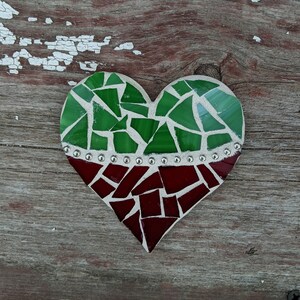 May include: A heart-shaped mosaic art piece. The top half features green glass pieces, the bottom half is red. A row of small silver-colored embellishments separates the two colors. The mosaic is set against a weathered wood background.