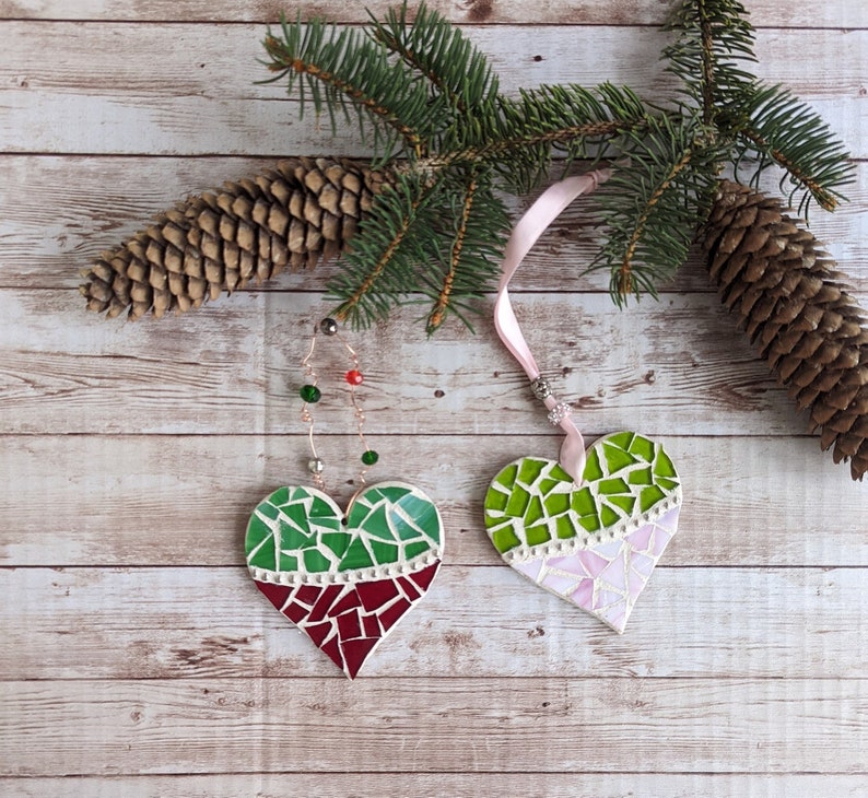 May include: Two heart-shaped mosaic ornaments with green and red, and green and pink designs, hang from a pink ribbon and a wire with beads. A pine branch and pine cone are in the background, set against a wood-grain surface.