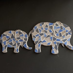 K&ouml;nnte beinhalten: Zwei Mosaik-Elefantenfiguren. Die Elefanten bestehen aus blauen, grauen und wei&szlig;en Fliesen. Der gr&ouml;&szlig;ere Elefant hat ein Perlendetail auf dem Kopf. Der Hintergrund ist schwarz.