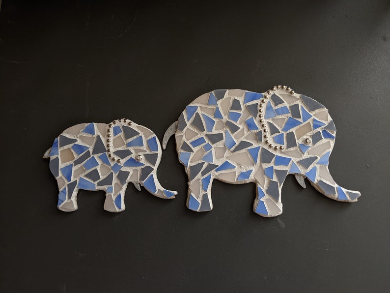 K&ouml;nnte beinhalten: Zwei Mosaik-Elefantenfiguren. Die Elefanten bestehen aus blauen, grauen und wei&szlig;en Fliesen. Der gr&ouml;&szlig;ere Elefant hat ein Perlendetail auf dem Kopf. Der Hintergrund ist schwarz.