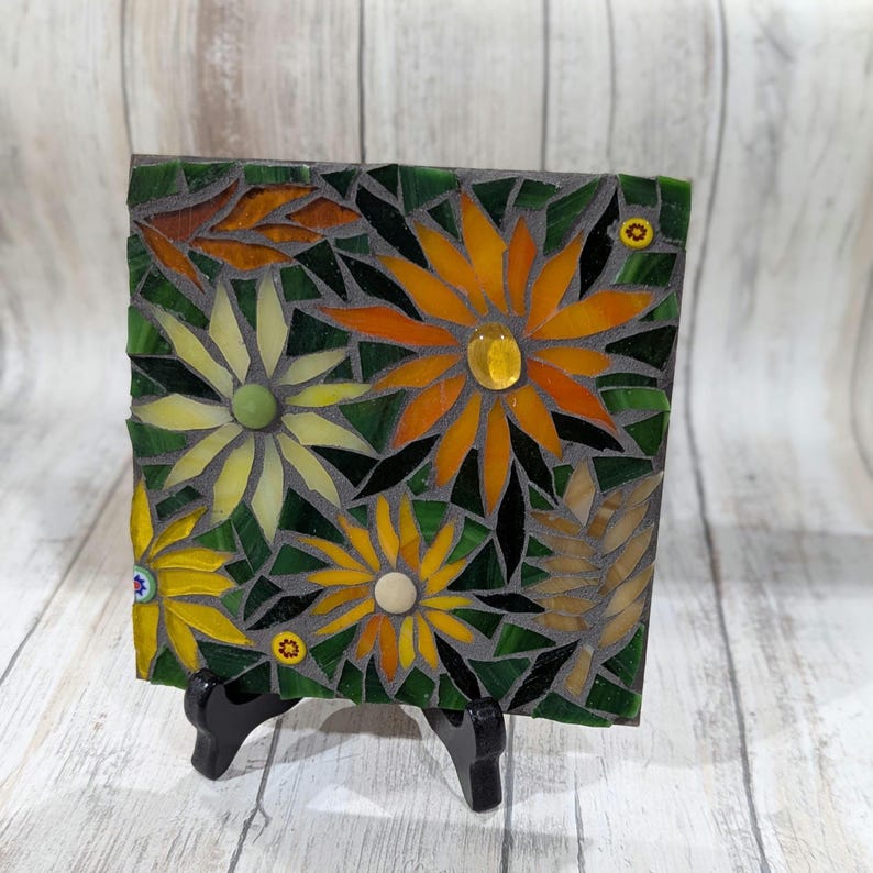 May include: Square mosaic art piece featuring a floral design with yellow, orange, and green glass tiles. The mosaic depicts stylized flowers with a dark gray background and is displayed on a black stand.