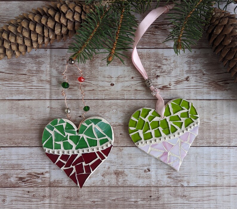 May include: Two heart-shaped mosaic ornaments. One features green and burgundy tiles, the other green and pink. Both have a white border and are suspended by ribbons. Festive holiday decor.