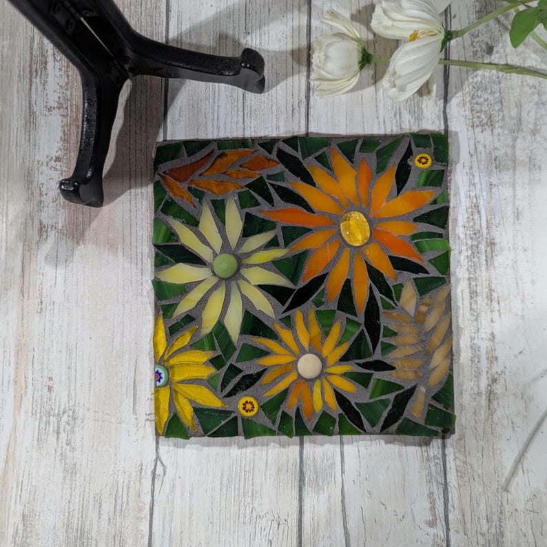 May include: A square mosaic tile featuring a floral design with yellow and orange flowers, green leaves, and a gray background. The mosaic is made of small, colorful glass pieces. The tile is a decorative item.