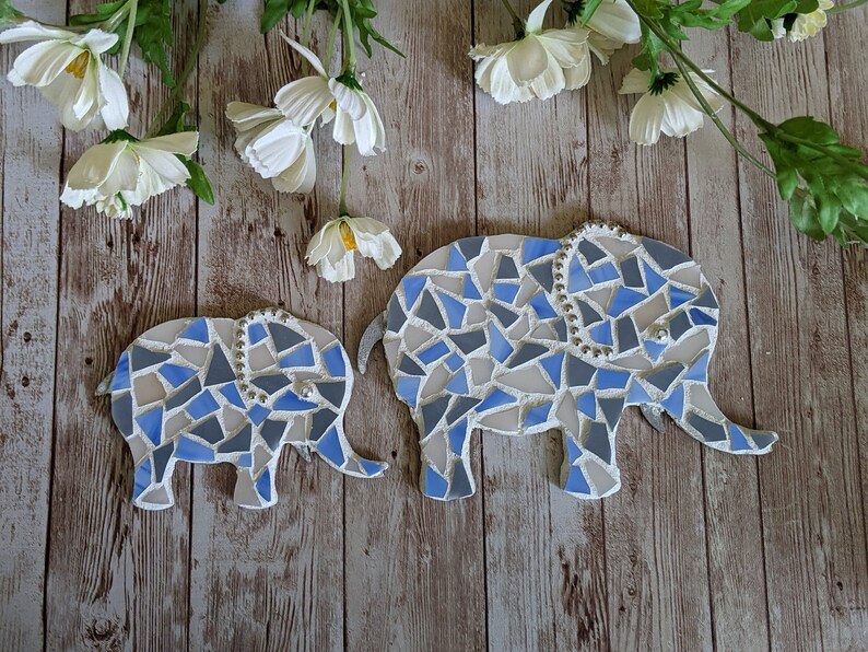 K&ouml;nnte beinhalten: Zwei Mosaik-Elefantenfiguren in Blau, Grau und Wei&szlig; mit einem Perlenrand. Die Elefanten befinden sich auf einer Holzoberfl&auml;che, im Hintergrund wei&szlig;e Blumen und Gr&uuml;n.