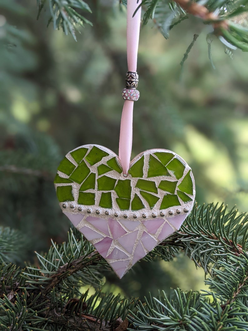 May include: A heart-shaped ornament with a mosaic design. The top half features green glass pieces, while the bottom half has pink glass. A pink ribbon and decorative beads are attached for hanging. The ornament is displayed against a backdrop of green pine needles.