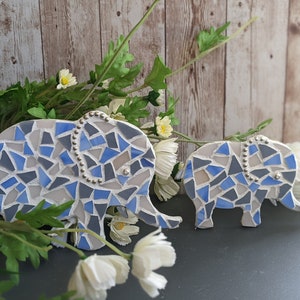 K&ouml;nnte beinhalten: Zwei Mosaik-Elefantenfiguren. Die Elefanten bestehen aus blauen, grauen und wei&szlig;en Fliesen. Der gr&ouml;&szlig;ere Elefant hat eine Perlenverzierung entlang seines R&uuml;ckens. Die Figuren werden mit wei&szlig;en Blumen und Gr&uuml;nzeug ausgestellt.