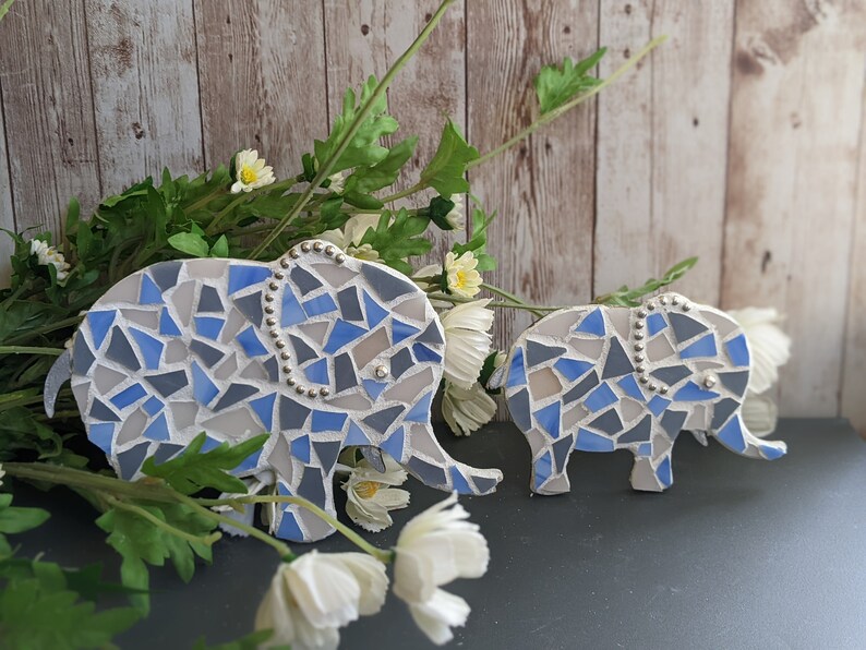 K&ouml;nnte beinhalten: Zwei Mosaik-Elefantenfiguren. Die Elefanten bestehen aus blauen, grauen und wei&szlig;en Fliesen. Der gr&ouml;&szlig;ere Elefant hat eine Perlenverzierung entlang seines R&uuml;ckens. Die Figuren werden mit wei&szlig;en Blumen und Gr&uuml;nzeug ausgestellt.