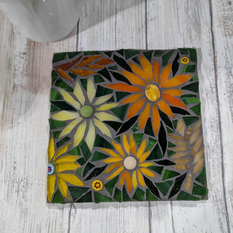 May include: Square mosaic coaster featuring a floral design. The mosaic is composed of various colored glass pieces, including yellow, orange, green, and brown. The flowers have yellow and orange petals with green and black leaves.