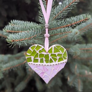 May include: A heart-shaped mosaic ornament with a pink ribbon for hanging. The top half of the heart is green, and the bottom half is pink. The ornament is decorated with small silver beads.