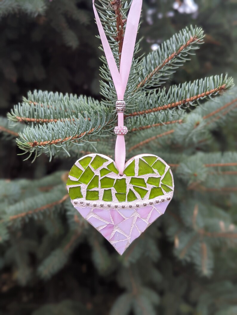 May include: A heart-shaped mosaic ornament with a pink ribbon for hanging. The top half of the heart is green, and the bottom half is pink. The ornament is decorated with small silver beads.