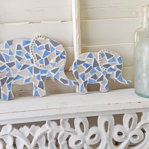 K&ouml;nnte beinhalten: Zwei Mosaik-Elefantenfiguren in Blau-, Grau- und Wei&szlig;t&ouml;nen. Die Elefanten haben Perlenakzente und werden auf einem wei&szlig;en Regal ausgestellt. Der gr&ouml;&szlig;ere Elefant ist etwa 20 cm lang, der kleinere etwa 13 cm.