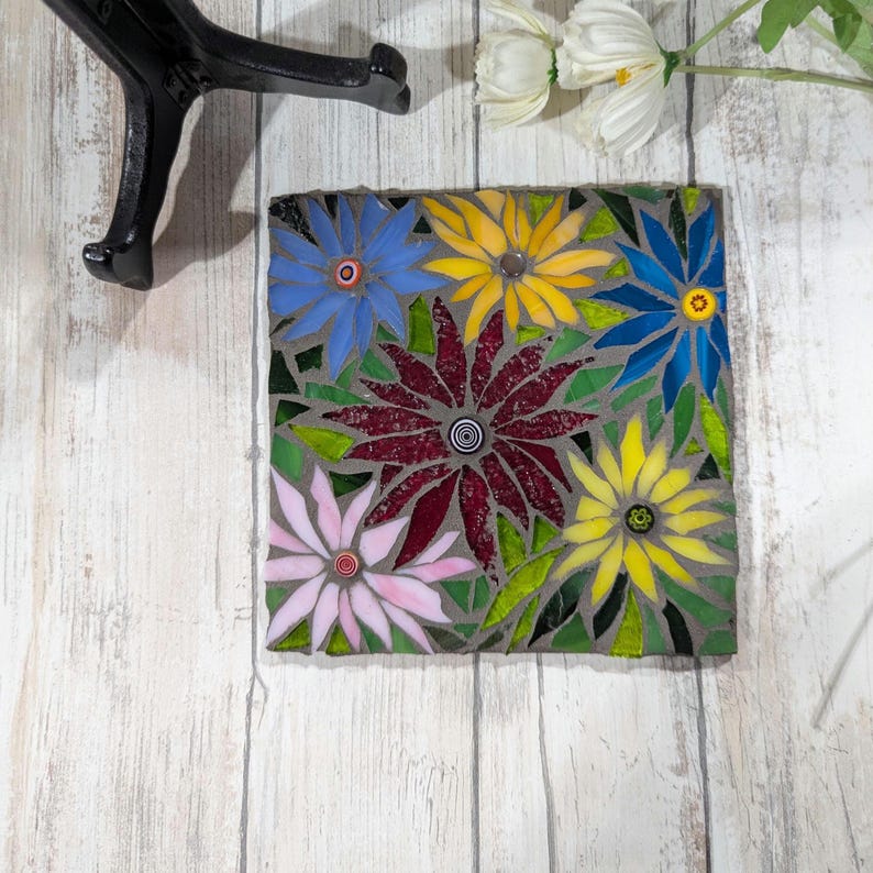 May include: A square mosaic tile featuring a floral design with blue, yellow, pink, and burgundy flowers. The flowers have detailed centers and are set against a gray background with green leaves. The tile is on a white wooden surface.
