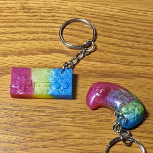 May include: Two keychains with silver rings and chains. One is a rectangular shape with a controller design in pink, yellow, and blue. The other is a curved shape in pink, purple, green, and blue.